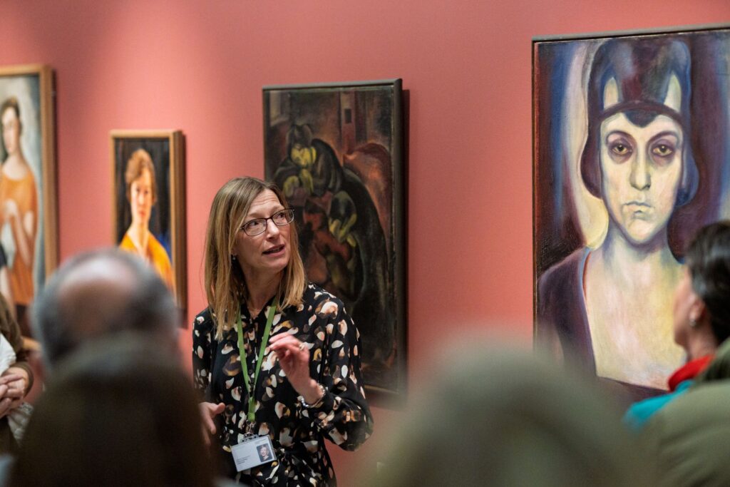 Docent with glasses and a green lanyard speaks to a museum group beside colorful paintings on a pink wall.