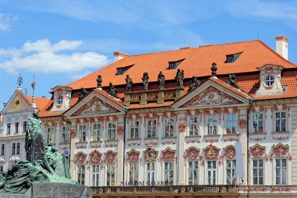 Facade of an ornate Baroque-style building with pink reliefs and an orange tiled roof, statues along the roofline, and a bronze monument in the foreground.