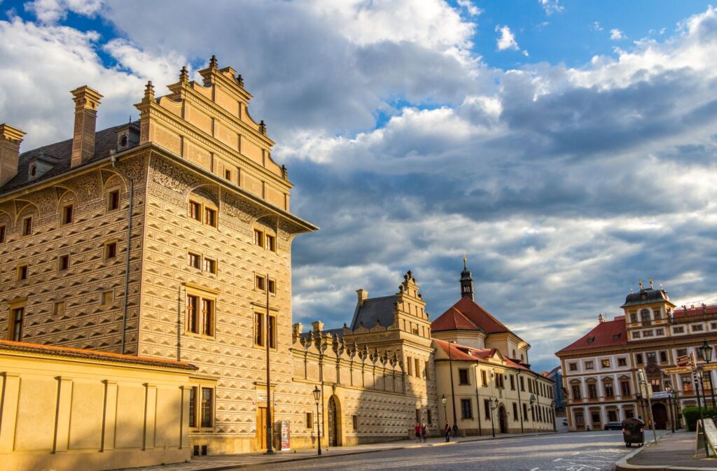Historic European street with an ornate, patterned stone building and red-tiled roofs under a blue sky with clouds.