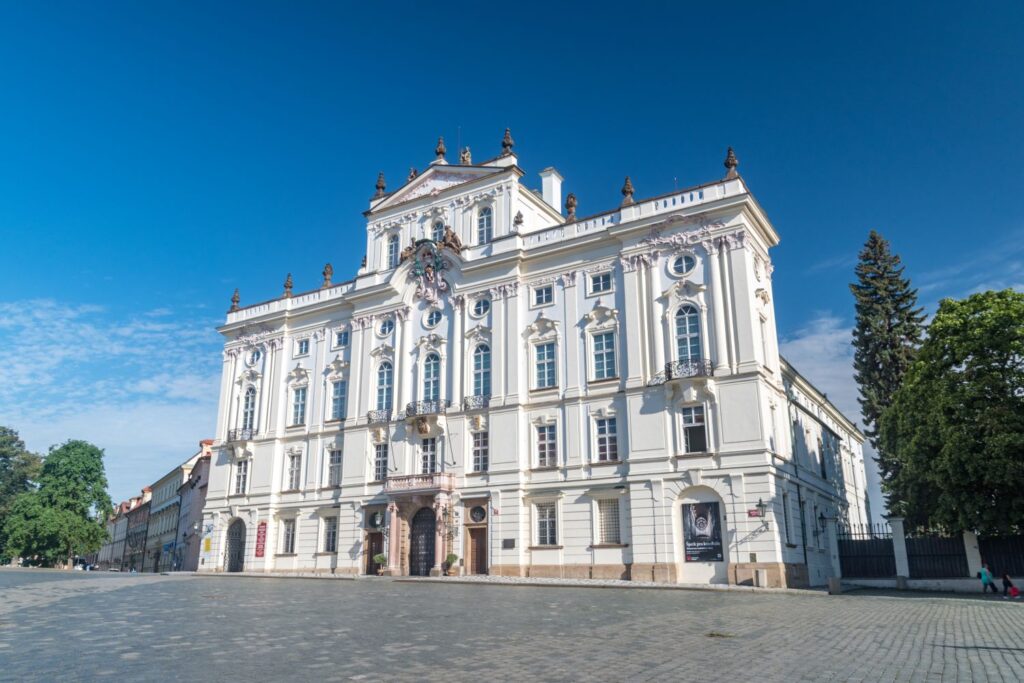 White ornate historic building with sculpted details and arched windows on a cobblestone square under a bright blue sky.