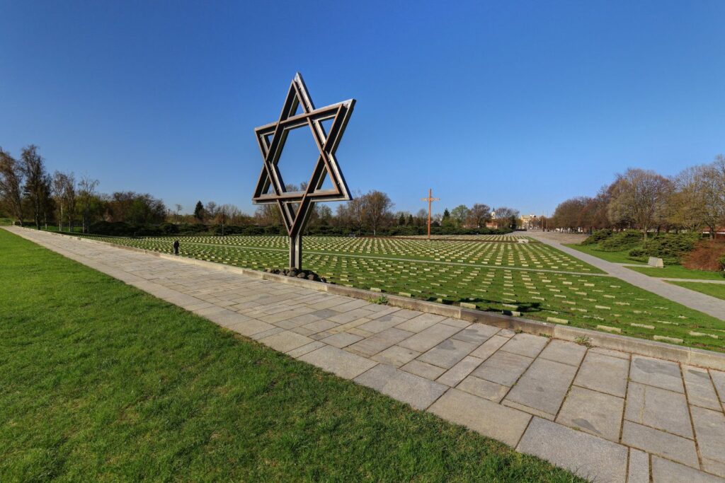 Wide-angle view of a memorial park featuring a tall metal Star of David sculpture and rows of small gravestones across the lawn under a clear blue sky.