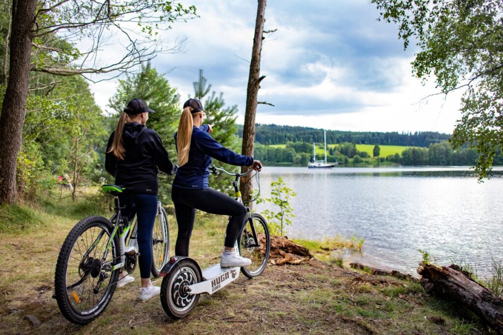 Two women in athletic clothes with bikes on a forested lakeside path, one standing on a white scooter attached to a bike, pointing toward the water.